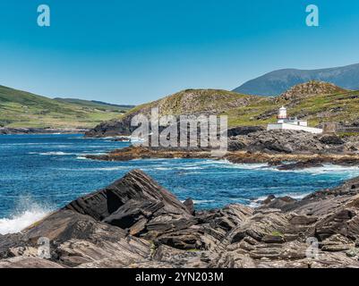 Ein atemberaubender Blick auf die Küste mit einem Leuchtturm auf zerklüfteten Felsen, umgeben von klarem blauem Wasser und sanften Hügeln. Ideal für Naturthemen, Stockfoto