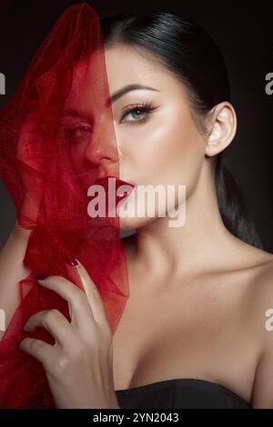 Die Frau steht in einem Studio mit dunklem Hintergrund und hält einen durchsichtigen roten Stoff vor ihr Gesicht. Sie zeigt leuchtend rote Lippen und fesselndes Make-up Stockfoto