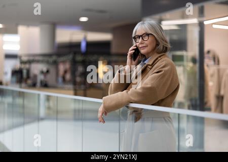 Eine reife Frau, die eine Brille trägt, spricht tagsüber am Telefon, während sie sich in einem modernen Einkaufszentrum gegen ein Geländer lehnt Stockfoto