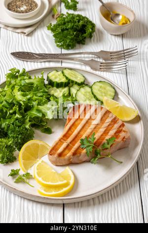 Gegrilltes Thunfischsteak mit frischem Salat, Gurkenscheiben und Zitronenscheiben auf einem Teller auf weißem Holztisch mit Gabeln, gelbem Senf und gemahlenem schwarzem Pfeffer Stockfoto