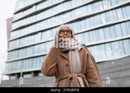 Eine Frau in einem schicken Mantel spricht an einem kühlen Tag vor einem modernen Bürogebäude Stockfoto