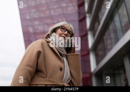 Eine ältere Frau in einem braunen Mantel spricht an einem bewölkten Tag in der Nähe eines modernen Gebäudes in einer Stadt auf ihrem Smartphone Stockfoto