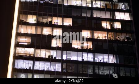 Bürogebäude am Abend mit beleuchteten Arbeitsplätzen. Moderner, verglaster Wolkenkratzer im Finanzviertel. Späte Öffnungszeiten im Büro. Stockfoto