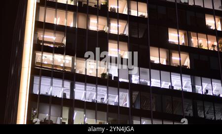 Bürogebäude am Abend mit beleuchteten Arbeitsplätzen. Moderner, verglaster Wolkenkratzer im Finanzviertel. Späte Öffnungszeiten im Büro. Stockfoto