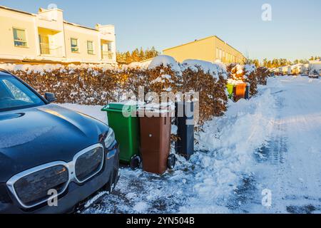 Schneebedeckte Wohnstraße mit geparktem schwarzen Auto, grünen und braunen Mülltonnen und Hecken am Wintertag. Stockfoto