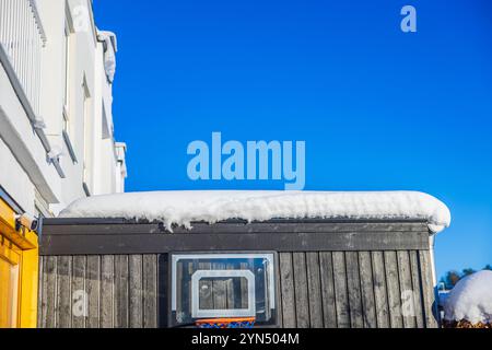 Basketballkorb an dunkler Holzwand unter dem schneebedeckten Dach vor dem leuchtend blauen Winterhimmel. Stockfoto