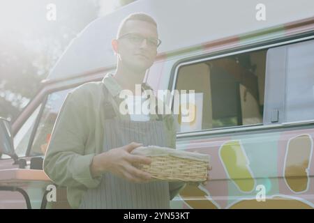 Porträt eines jungen kaukasischen Food-Truck-Arbeiters, der stillsteht, mit Korbkästen in Händen, beleuchtet von warmen Sonnenstrahlen Stockfoto