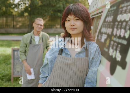 Mittelgroßes Nahporträt einer asiatischen Arbeiterin, die in die Kamera blickt und lächelt, während sich ihre Kollegin auf einen Esstisch lehnt, in einem unscharfen Hintergrund Stockfoto