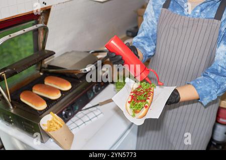 Hochwinkelaufnahme einer nicht erkennbaren Küchenchefin, die Ketchup in Hot Dog hinzufügt, während sie neben dem Grill mit Brötchen steht Stockfoto