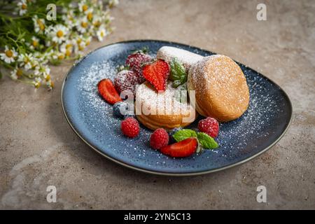 Flauschige japanische Souffelpfannkuchen, mit Puderzucker bestäubt, serviert auf einem blauen Teller mit frischen Beeren, Minzblättern und Kamillenblüten Stockfoto