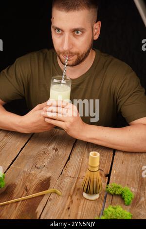Ein nachdenklicher Mann genießt ein Glas cremigen Matcha Latte mit einem Bambusquirl auf einem Holztisch. Ideal für Themen wie Achtsamkeit, Wellness und Tee-Kultu Stockfoto