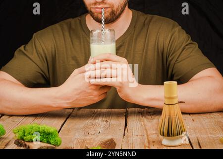 Ein nachdenklicher Mann genießt ein Glas cremigen Matcha Latte mit einem Bambusquirl auf einem Holztisch. Ideal für Themen wie Achtsamkeit, Wellness und Tee-Kultu Stockfoto
