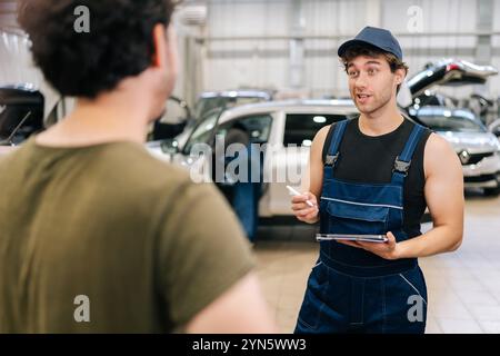 Männlicher Garagenarbeiter in Uniform und nicht erkennbarer Mann Kunde spricht, um Details von Autoreparaturen zu besprechen. Automatischer Mechaniker hält Tablet und Schreibvorgänge Stockfoto