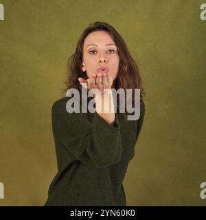 Lifestyles-Konzept. Eine Frau in einem grünen Pullover spritzt einen Kuss vor einem olivgrünen Hintergrund Stockfoto