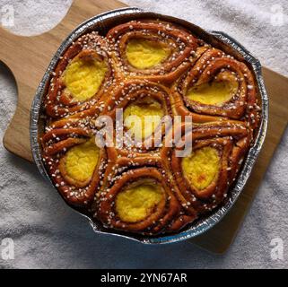 Süße Brötchen mit einem reichen Zimtgeschmack und einer herrlichen gelben Füllung sitzen in einer Metalldose auf einem Holzbrett. Die Brötchen sind mit Sesamsamen A belegt Stockfoto