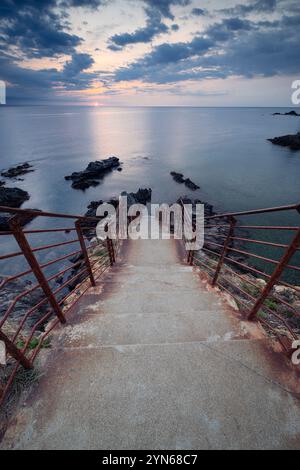 Sonnenaufgang über einer Bucht mit zerklüfteten Felsen, Betontreppe und rostigem Geländer an der Mittelmeerküste in der Nähe von Collioure, Cote Vermeille, Okzitanien Stockfoto