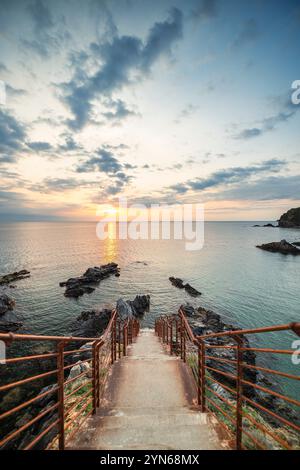 Sonnenaufgang über einer Bucht mit zerklüfteten Felsen, Betontreppe und rostigem Geländer an der Mittelmeerküste in der Nähe von Collioure, Cote Vermeille, Okzitanien Stockfoto