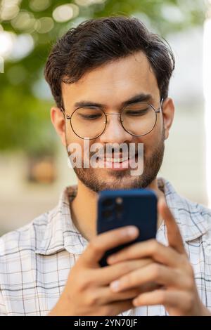 Indischer junger Mann, der per Smartphone Textnachrichten in sozialen Medien online schreibt, im Internet surft, sich entspannt, eine Pause im Freien macht. Ein Araber steht auf der Stadtstraße. Vertikal Stockfoto