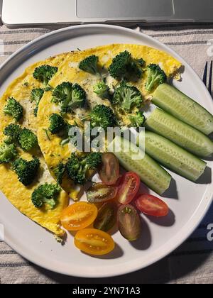 Zwei flauschige Omeletts mit Brokkoli werden auf einem Teller serviert, zusammen mit frischen Gurkenscheiben und bunten Kirschtomaten. Ein nahrhaftes Frühstück Stockfoto