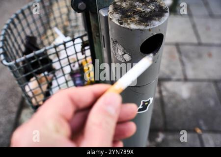 Augsburg, Bayern, Deutschland - 24. November 2024: Ein Mann hält eine brennende Zigarette vor einem öffentlichen Aschenbecher, Symbolbild für Rauchen im Freien und Tabakkonsum *** ein Mann hält eine brennende Zigarette vor einem öffentlichen Aschenbecher, Symbolbild für Rauchen im Freien und Tabakkonsum Stockfoto