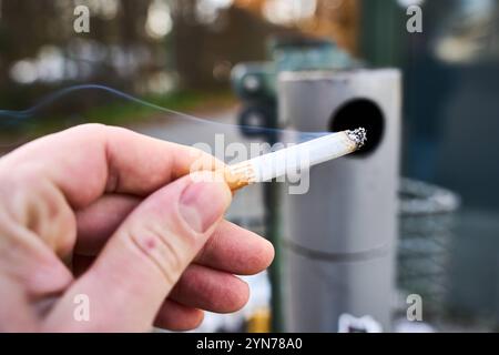 Augsburg, Bayern, Deutschland - 24. November 2024: Ein Mann hält eine brennende Zigarette vor einem öffentlichen Aschenbecher, Symbolbild für Rauchen im Freien und Tabakkonsum *** ein Mann hält eine brennende Zigarette vor einem öffentlichen Aschenbecher, Symbolbild für Rauchen im Freien und Tabakkonsum Stockfoto