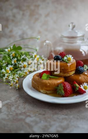 Flauschige japanische Pfannkuchen, serviert mit frischen Beeren, Minzblättern und Sirup, begleitet von Kamillenblüten und einer Teekanne aus Glas Stockfoto