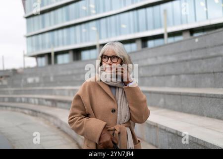 Eine ältere Frau mit Brille und braunem Mantel spricht am Telefon, während sie an einem bewölkten Tag vor einem Stadtgebäude steht Stockfoto
