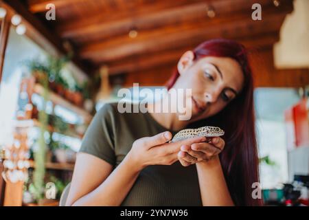 Junge Frau, die sanft eine Eidechse in den Händen hält, während sie in einem gemütlichen Innenraum mit viel Grün und Holzdekor sitzt Stockfoto