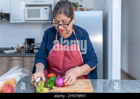 Eine Frau schneidet Gemüse auf einem Schneidebrett. Sie trägt eine rote Schürze und trägt eine Brille Stockfoto