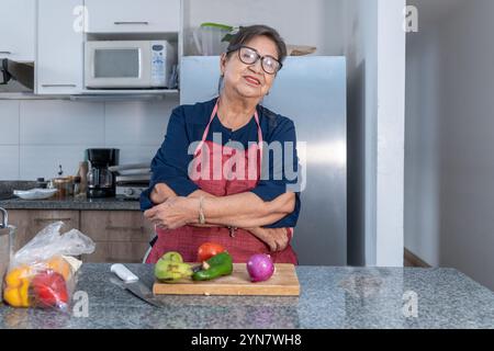 Porträt einer älteren Frau mit Brille, blauer Bluse und roter Schürze, die in der Küche lächelt, mit Gemüse, Tomaten, Zwiebeln und Pfeffer auf einem Schneidebrett Stockfoto