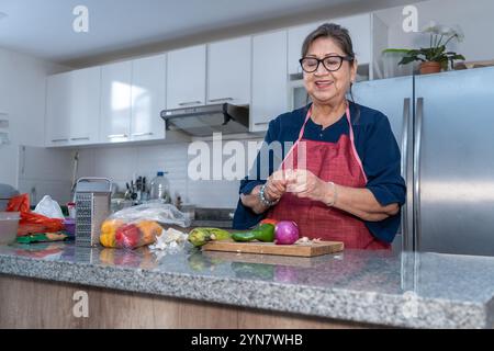 Senior Latina Frau schält Knoblauch in der Küche neben Gemüse wie Zwiebeln, Paprika und Tomaten. Die Frau lächelt beim Kochen Stockfoto