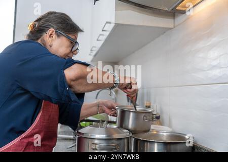 Eine Frau kocht in einer Küche mit einem Topf auf dem Herd. Sie trägt eine blaue Schürze und hat einen Löffel in der Hand. Eine Frau kocht in einer Küche mit Stockfoto