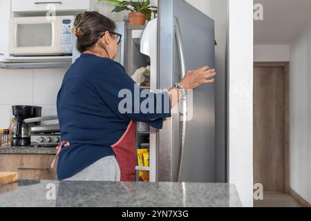 Eine Frau öffnet eine Kühlschranktür. Sie trägt ein blaues Hemd und eine rote Schürze Stockfoto