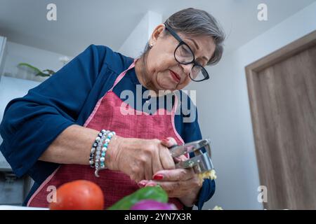Niedrigwinkelaufnahme einer älteren Latina, die Knoblauch mit einem Küchengerät zerquetscht. Die Frau trägt eine Brille, eine blaue Bluse und eine rote Schürze. Stockfoto