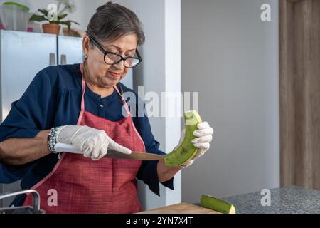 Eine Frau schneidet eine Banane mit einem Messer. Sie trägt eine Schürze und Handschuhe. Die Banane ist grün und wird halbiert Stockfoto