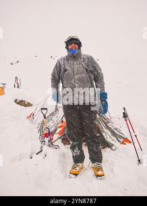Mann in einer schneebedeckten Berglandschaft, stehend vor seinem Zelt während eines Schneefalls, in Mendoza, Argentinien. Stockfoto