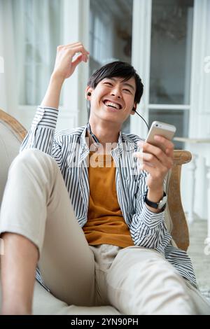 Ein Mann in seinen 20ern sitzt glücklich auf einem Sofa und schaut auf sein Smartphone Stockfoto