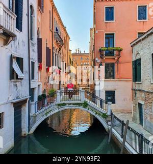 Eine Person entspannt sich auf einer malerischen Brücke, umgeben von bunten Gebäuden, die sich im ruhigen Wasser eines Kanals von Venedig spiegeln. Das warme Licht schafft ein magisches Ambiente in der Abenddämmerung. Stockfoto