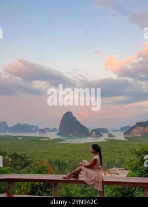 Eine Frau sitzt friedlich und blickt auf die atemberaubende, weitläufige Aussicht auf Kalksteininseln im Grünen, während die Sonne untergeht, und malt den Himmel in sanften Rosa- und Orangetönen in Phangnga Bay Thailand Stockfoto