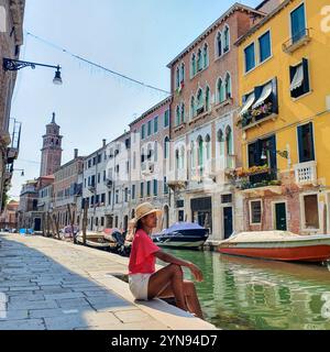 An einem sonnigen Nachmittag in Venedig sitzt eine Frau friedlich an einem ruhigen Kanal, umgeben von bezaubernden, farbenfrohen Gebäuden und sanft schwimmenden Booten, und genießt die Schönheit der Stadt. Stockfoto
