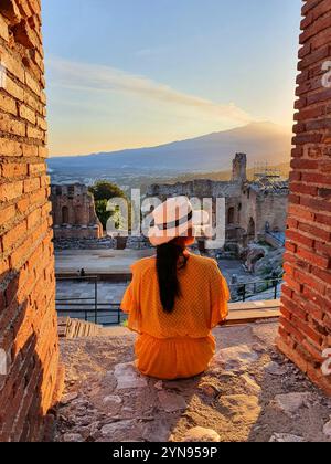 Eine Frau in stilvollem gelbem Kleid und breiter Krempe sitzt ruhig zwischen antiken Ruinen, der Himmel leuchtet sanft wie die Silhouette des Ätna, Taormin Stockfoto