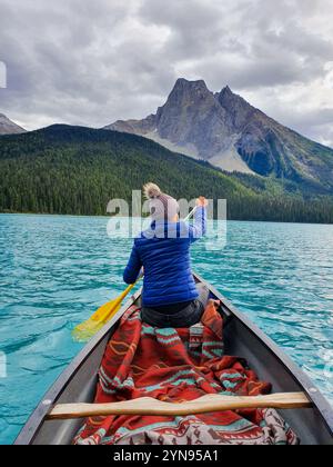 Eine Person in einer blauen Jacke paddelt mit einem Kanu auf türkisfarbenem Wasser, umgeben von majestätischen Bergen und üppigem Wald. Emerald Lake Yoho National Park Kanada Britisch Kolumbien Stockfoto