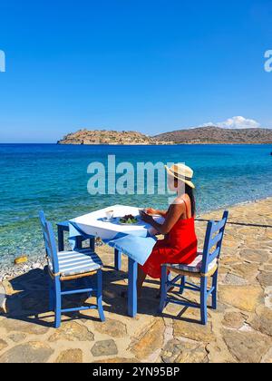 Unter der strahlenden Sonne schmeckt eine Frau in einem fließenden roten Kleid ihr Mittagessen an einem Tisch am Meer. Mit einem atemberaubenden Meerblick und sanften Wellen strahlt die Atmosphäre Ruhe und Schönheit aus. Kreta Griechenland Stockfoto