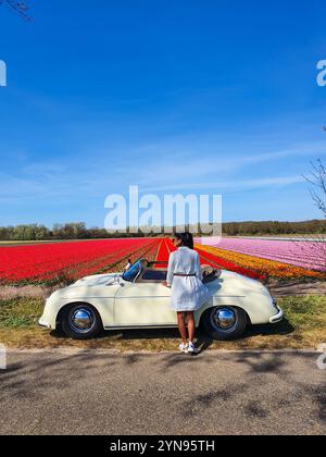 Unter einem leuchtend blauen Himmel steht eine Frau neben einem klassischen Cabrio und bestaunt den atemberaubenden Anblick lebendiger Tulpenfelder in den Niederlanden Stockfoto