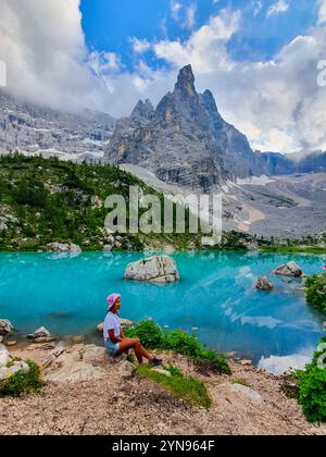 Eine Person sitzt friedlich an einem markanten türkisfarbenen See, umgeben von majestätischen Bergen und üppigem Grün. Die Wolken und das Sonnenlicht schaffen eine fesselnde Atmosphäre Lago Di Sorapis Dolomiten Italien Stockfoto