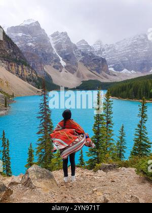Eine Person steht in eine farbenfrohe Decke gehüllt und blickt auf das atemberaubende türkisfarbene Wasser des Sees, eingerahmt von majestätischen Bergen und üppigem Grün. Moraine Lake Canada Stockfoto