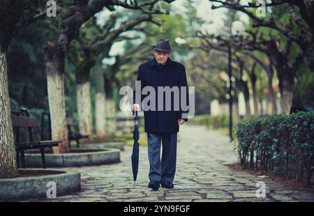 Ein älterer Mann mit dunklem Mantel und Hut schlendert durch einen ruhigen Park, der von Bäumen gesäumt ist. Er trägt einen Schirm, während er auf einem Steinweg unter einem läuft Stockfoto