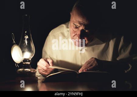 Ein älterer Mann sitzt an einem Holztisch, vertieft in ein Buch, das von einer flackernden Laterne beleuchtet wird. Das gedämpfte Licht schafft eine warme und intime Atmosphäre Stockfoto