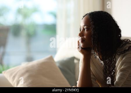 Eine zufriedene schwarze Frau, die durch ein Fenster nachdenkt und zu Hause auf einer Couch sitzt Stockfoto