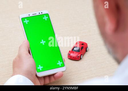 Mann, der ein leeres Telefon mit grünem Bildschirm in vertikaler Position neben dem roten Spielzeugauto hält. Anwendung zum Verkauf und Mieten von Autos, Online-Versicherung kaufen. Stockfoto